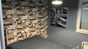 Une chambre avec un lit en métal, une couvre-lit grise, un mur décoré d’un motif imitant des briques et une fenêtre avec des stores.