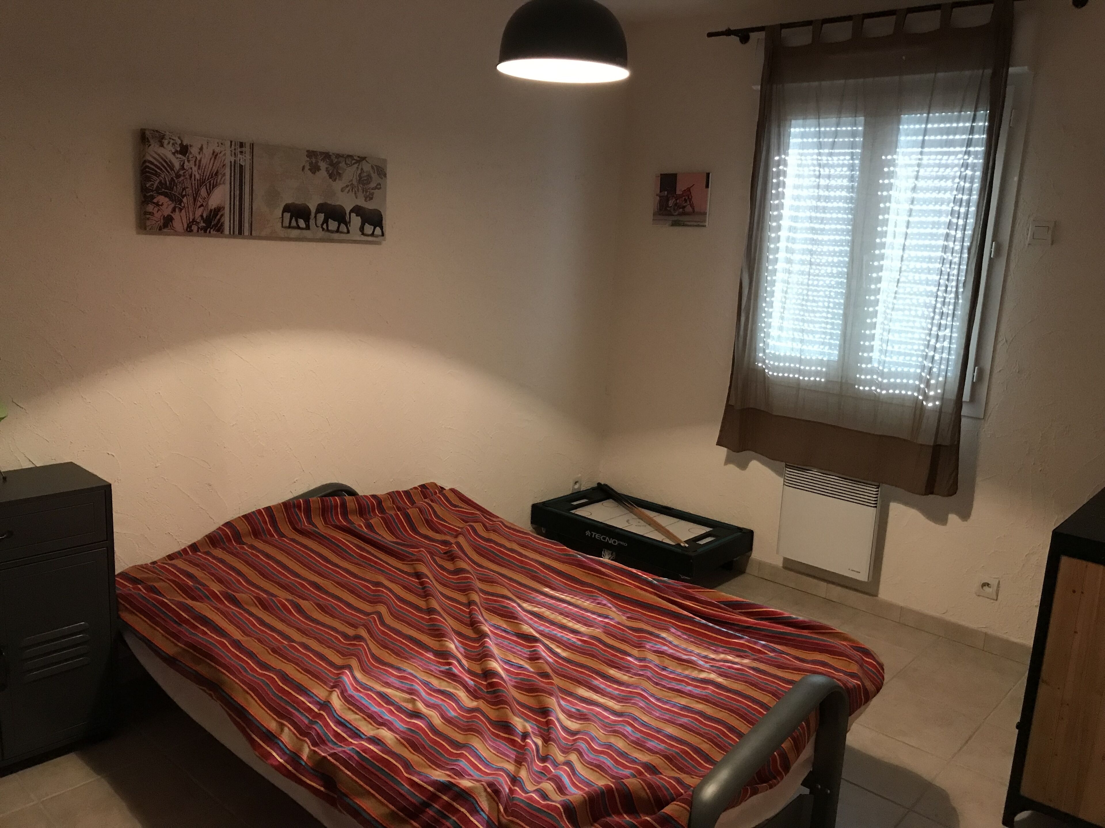 Ein kleiner, schlicht eingerichteter Raum mit Bett, Schreibtisch, Heizkörper und einem Fenster mit Vorhängen.