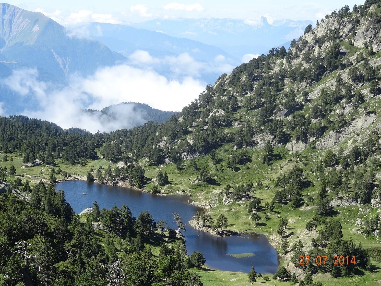 A mountainous landscape with a lake surrounded by dense forests and rocky terrain.