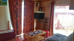 A cozy living room with a wooden ceiling, a television on a wooden stand, a red ottoman, a quilted sofa, and a window with curtains. - CHALET DES CIMES CHAMROUSSE / 3* TOURIST HOUSE / NEAR THE SLOPES (Chamrousse)