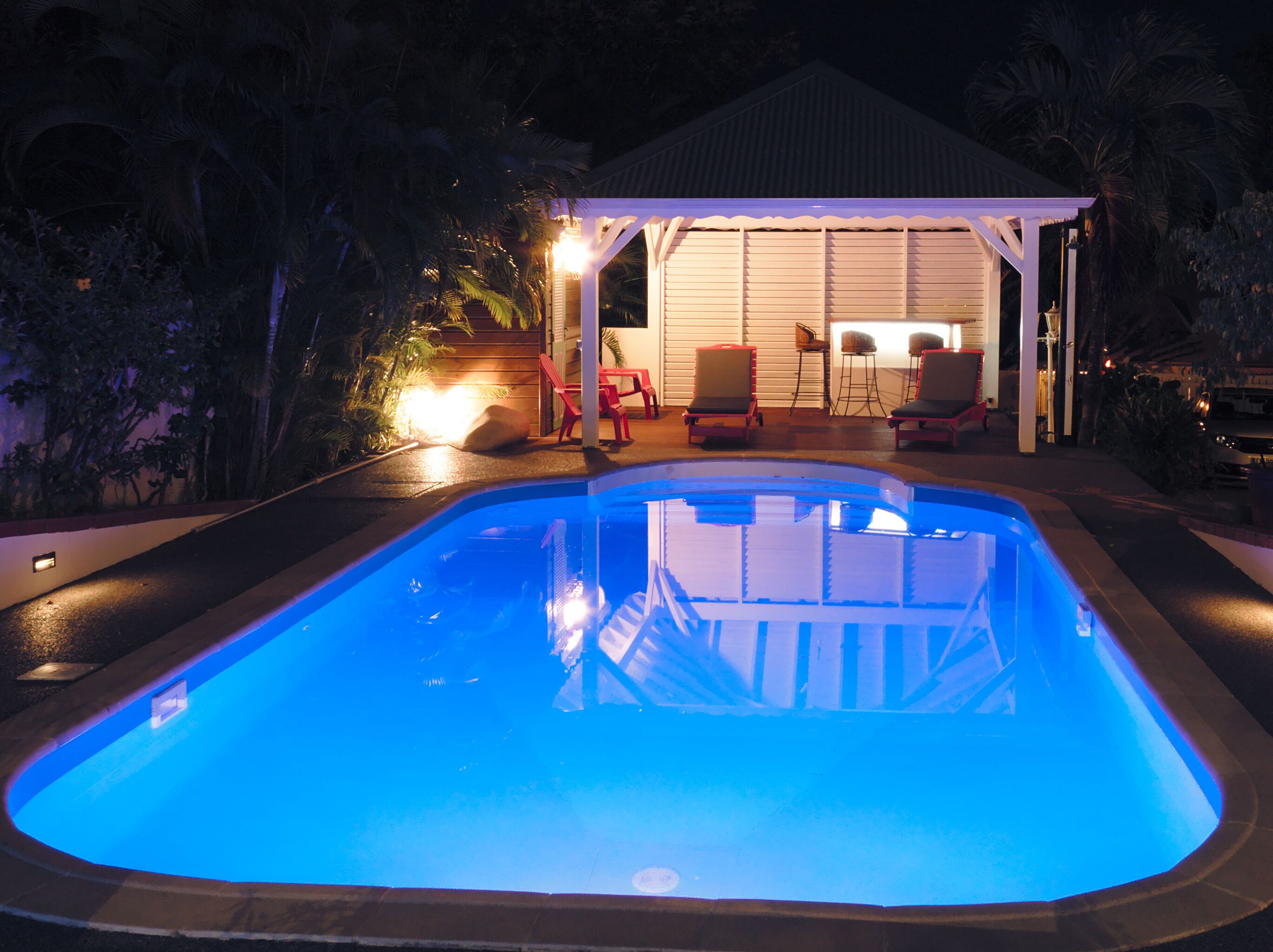 A swimming pool with a covered patio area, red chairs, and a small table.