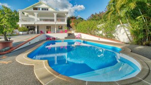 Une maison à deux étages avec une piscine, entourée de plantes tropicales et surplombant un ciel d’un bleu limpide.
