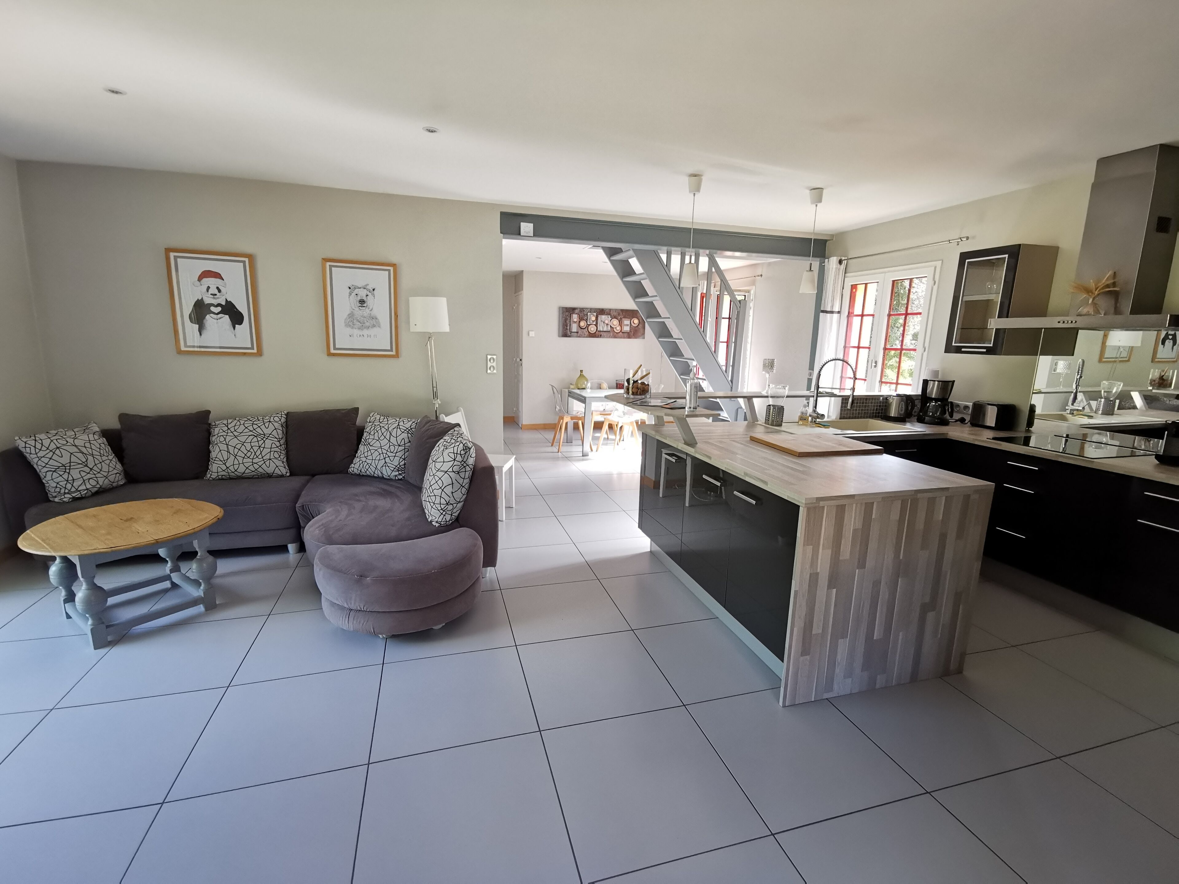 A modern living room with a sofa, ottoman, and coffee table, a kitchen with a central island, and a staircase leading to an upper level.