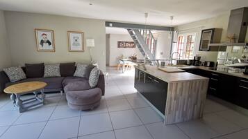 A modern living room with a sofa, ottoman, and coffee table, a kitchen with a central island, and a staircase leading to an upper level.