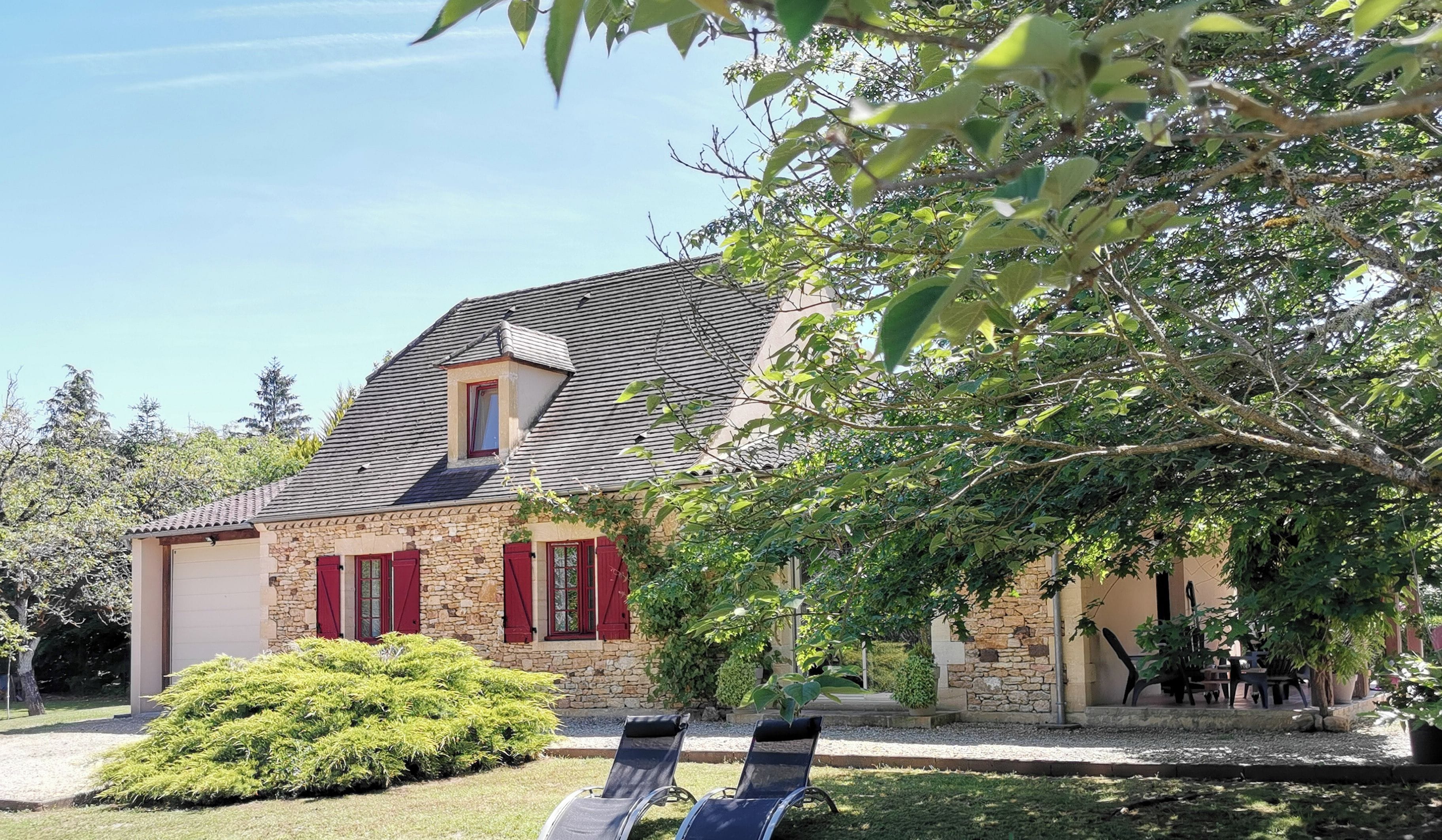 A stone house with a sloped roof, red shutters, and a small attached garage. There are two lounge chairs in the front yard.