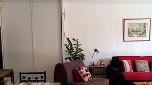 Een zithoek met een tafel en stoelen, een bank, een rode slaapbank, en een lijst met een afbeelding aan de muur.