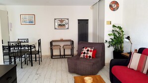 Een woonkamer met een bruine fauteuil, een rode bank, een houten salontafel, een eettafel met stoelen, een lijst met een afbeelding, een klok en een potplant.