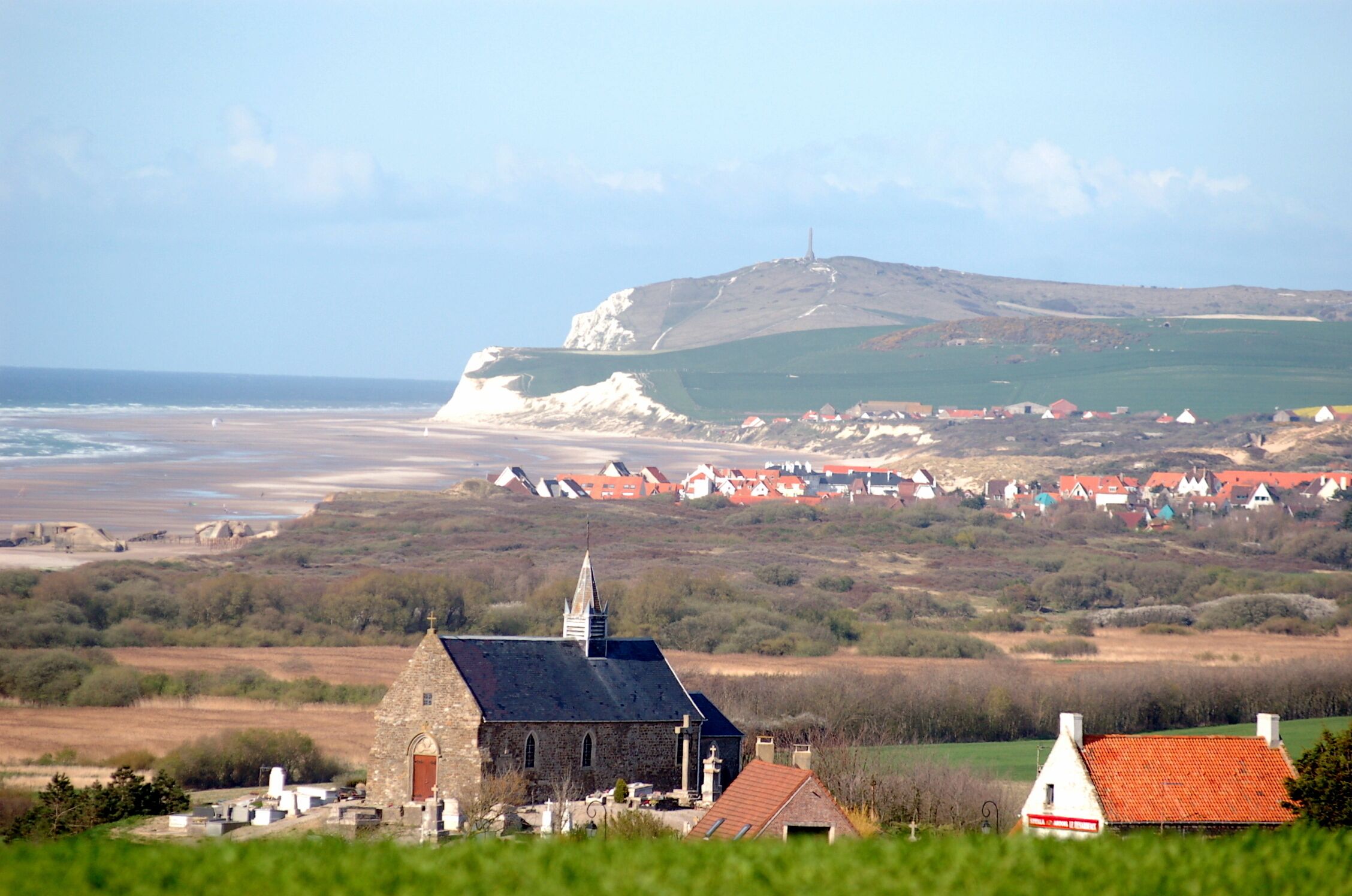 A coastal village with a prominent church, a hilltop monument, and a sea view.