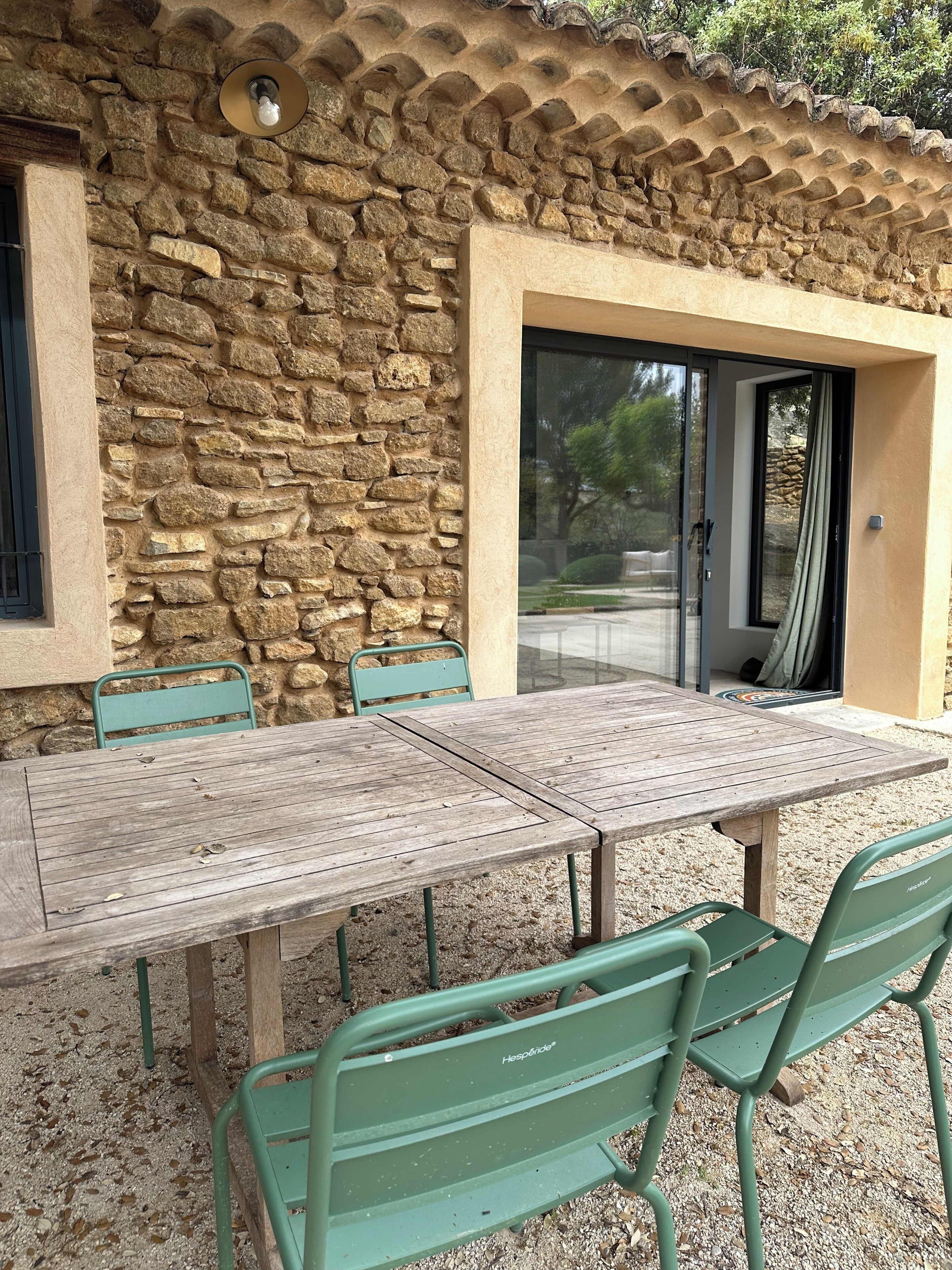 Un bâtiment en pierre avec une table en bois et des chaises vertes, une porte et une fenêtre.