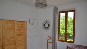A bedroom with a wooden wardrobe, a bed, a small table with a floral tablecloth, a wall-mounted mirror, and a window with a view of greenery. - TYPICAL VILLAGE HOUSE CORSE  (Levie)