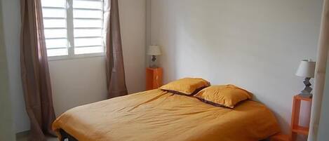 A bedroom with a bed, curtains, an air conditioning unit, and a nightstand with a lamp.