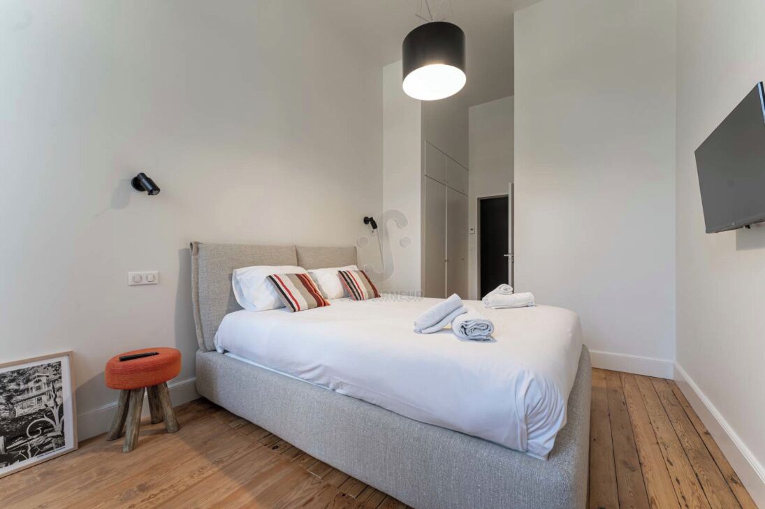 A modern bedroom with a bed, a small stool, a framed picture, and a TV on the wall.