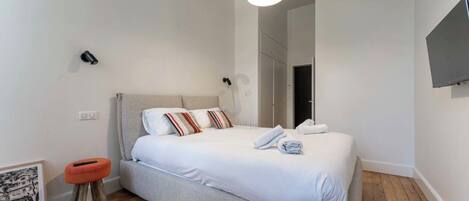 A modern bedroom with a bed, a small stool, a framed picture, and a TV on the wall.