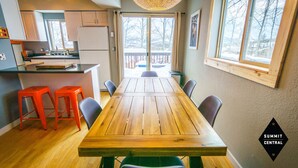A dining area with a wooden table and four chairs, a kitchen with a refrigerator and cabinets, and a view of a snowy deck through large windows. - Cozy, modern 2BD/2BA townhome minutes from 4 world class ski resorts. (Silverthorne)