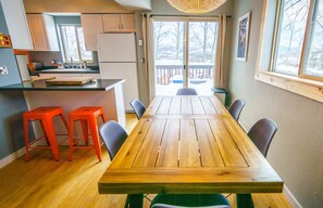 A dining area with a wooden table and four chairs, a kitchen with a refrigerator and cabinets, and a view of a snowy deck through large windows.