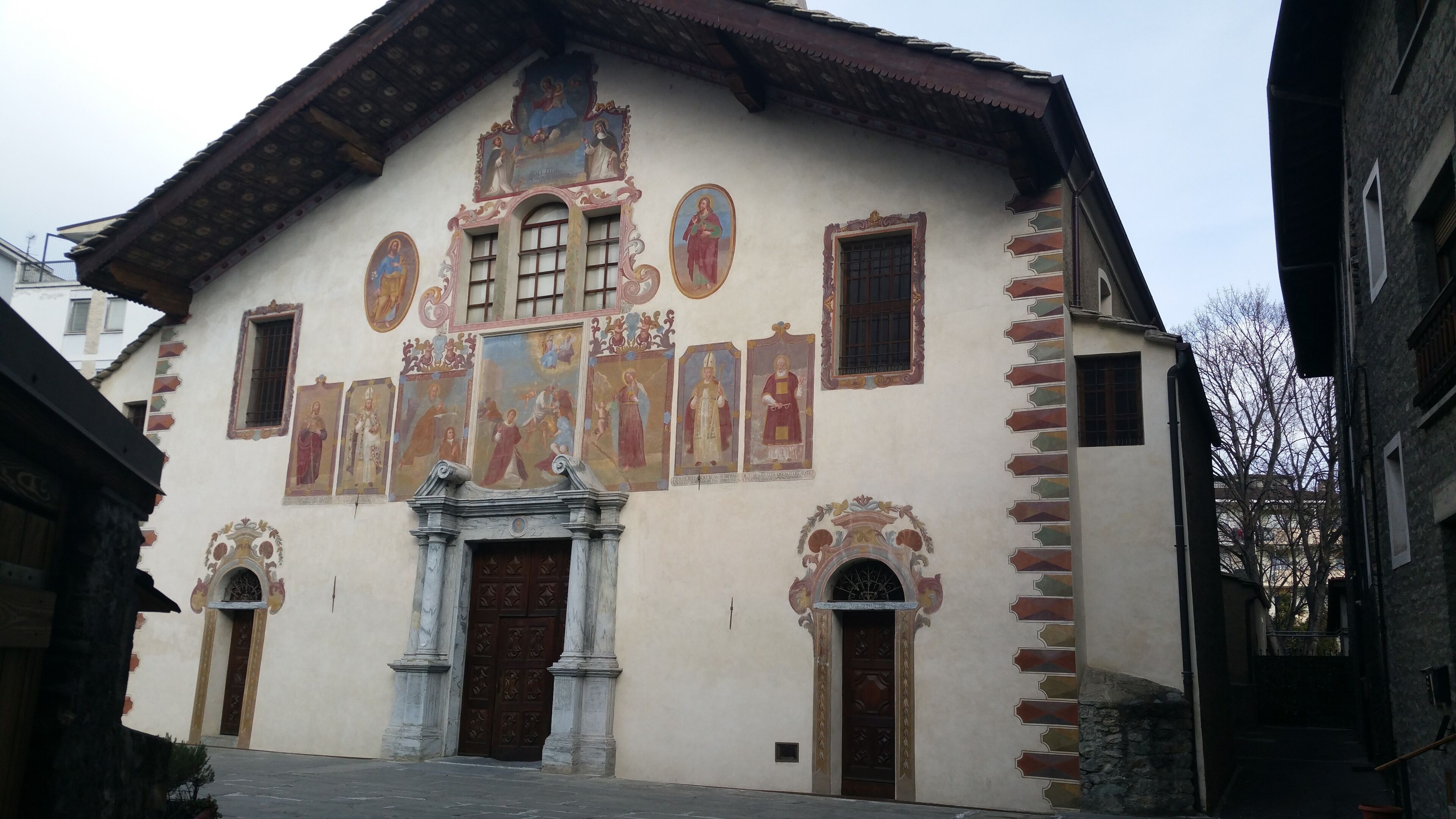 Un edificio con frescos religiosos, una entrada central con un arco decorativo y dos puertas laterales.