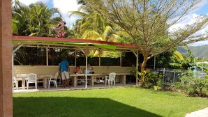 A covered outdoor seating area with a table and chairs, surrounded by lush greenery and a view of hills. - Nice apartment perfect also for people with reduced mobility (Sainte-Anne)