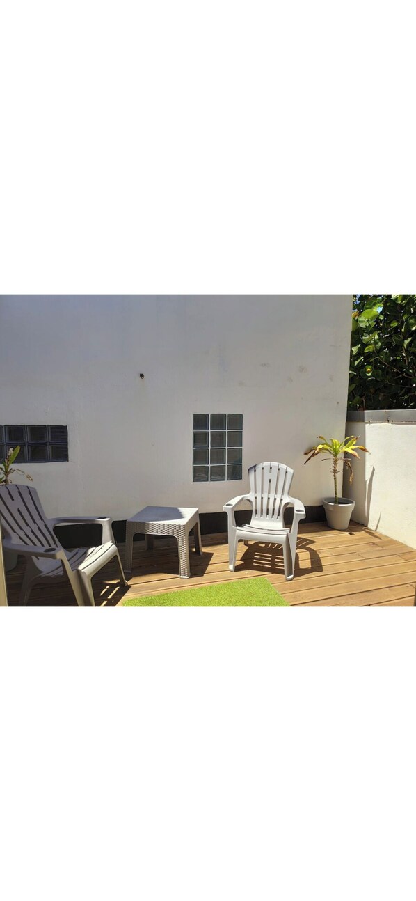 En uteplats med två vita plaststolar och ett litet bord, en krukväxt och en vit vägg med fönster.