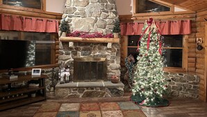 A cozy cabin interior with a stone fireplace, a decorated Christmas tree, and a wooden entertainment center. - Working Horse Ranch With Indoor Pool!! (Grantville)