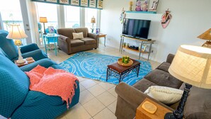 A living room with a blue patterned rug, a brown sofa, a blue armchair, and a television on a stand. - One NITE  MINIMUM  5 steps off deck onto beach; 10 steps out front door to pool. (Panama City Beach)