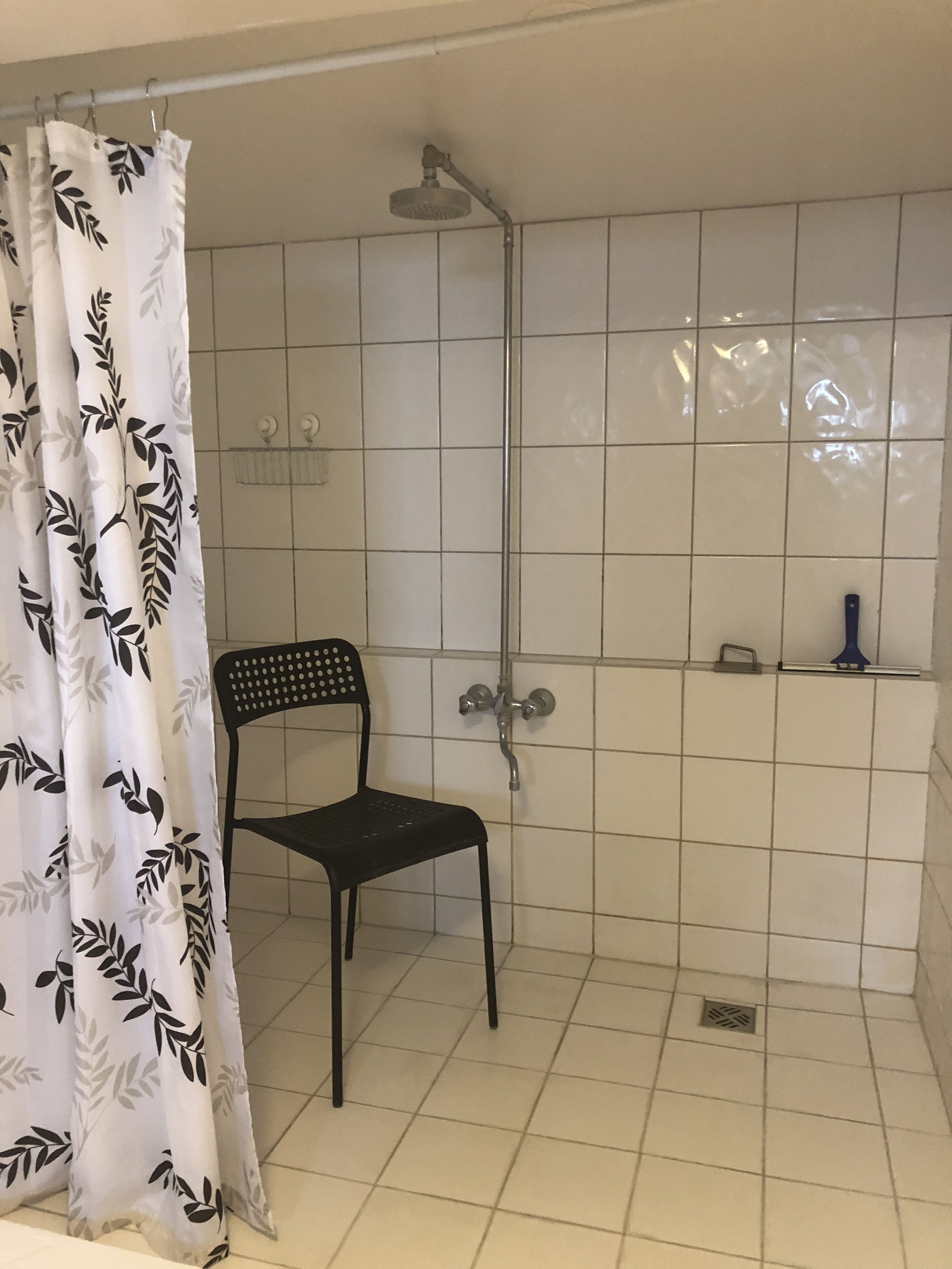 A bathroom with a shower, a chair, and a shelf with toiletries.
