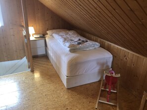 A single bed in a wooden room with a sloped ceiling, a nightstand, and a red chair.