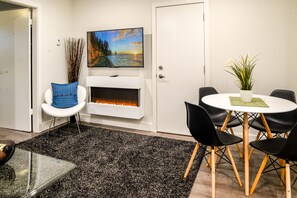 Una sala moderna con chimenea, un televisor montado encima, un comedor con una mesa redonda y sillas, y una planta decorativa.