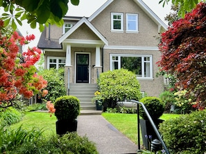 Une maison à deux étages avec un porche à l’avant, une porte noire et un petit jardin agrémenté de diverses plantes et fleurs.
