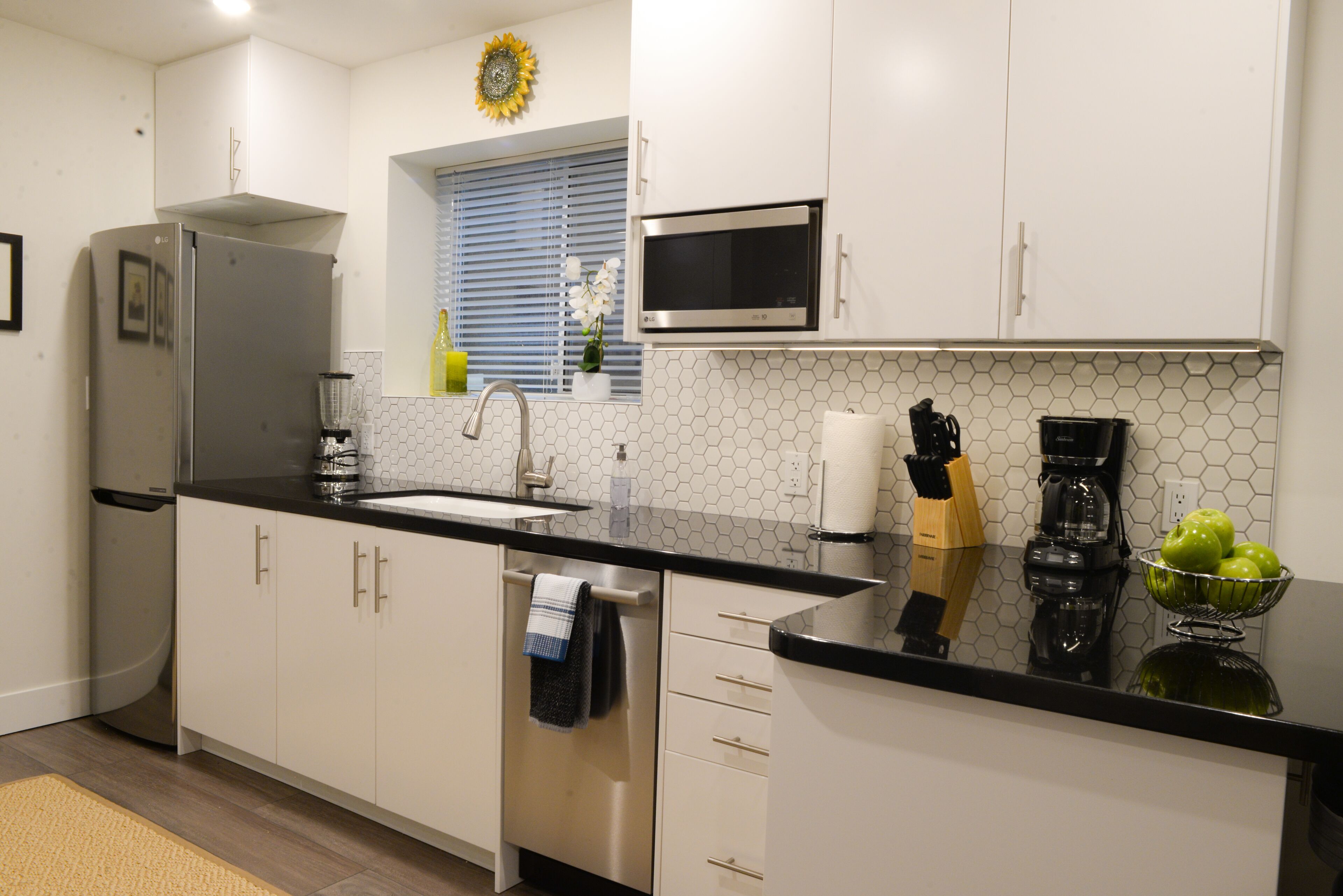 A modern kitchen with white cabinets, a stainless steel refrigerator, a black countertop, a sink, a microwave, and a coffee maker.