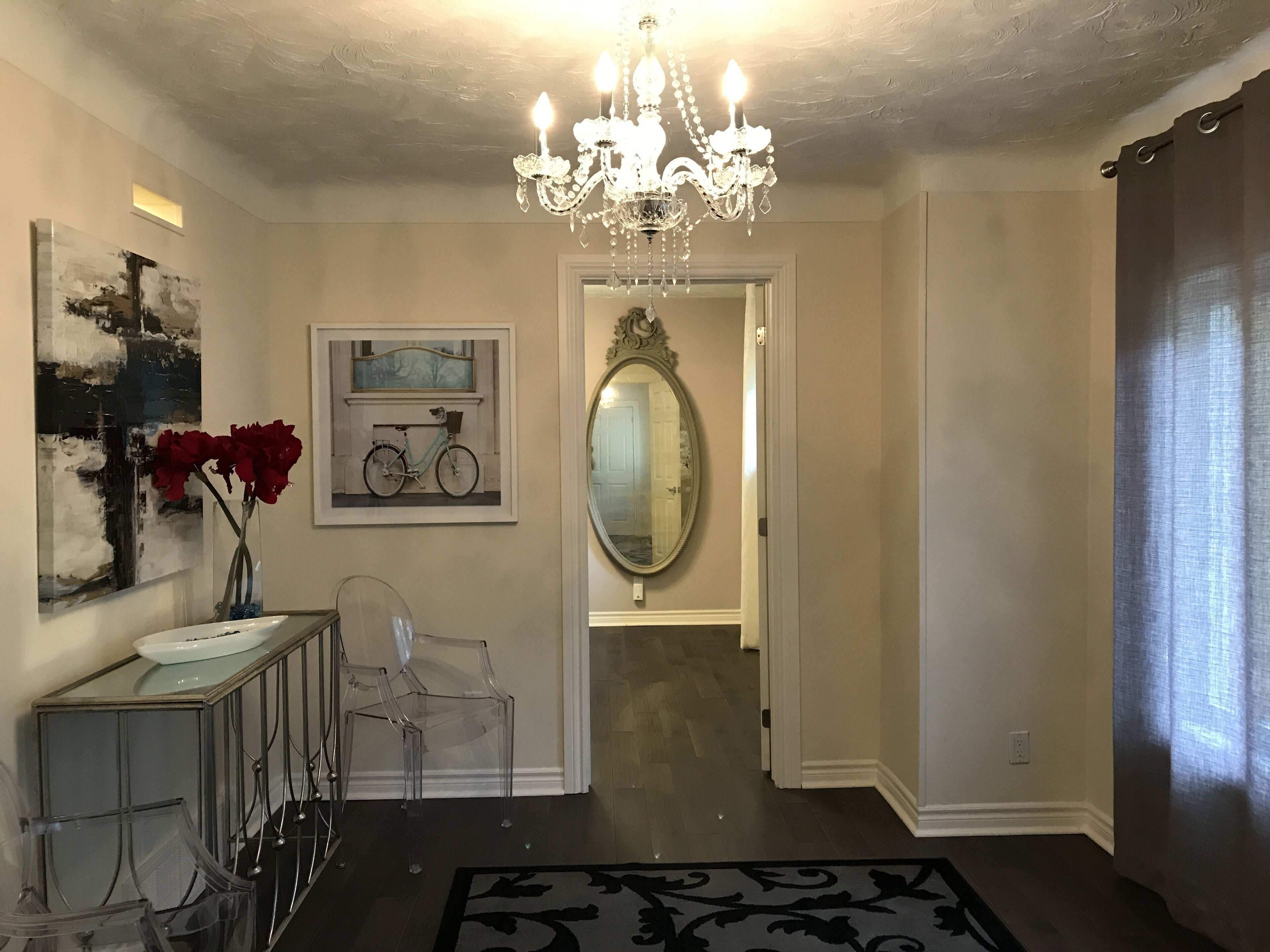 A hallway with a chandelier, a mirror, a bicycle painting, and a vase with red flowers.