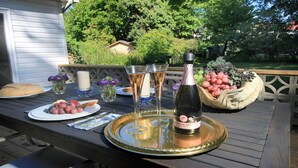 Une terrasse en bois avec une table dressée pour un repas, agrémentée d’une bouteille de vin, de verres et d’un panier de fruits.