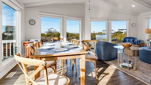 Un'ampia sala da pranzo con tavolo e sedie in legno, grandi finestre che si affacciano sul paesaggio e un'area salotto accogliente con un divano blu e un tavolino.