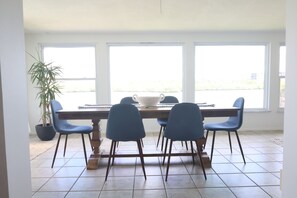 Zona pranzo con tavolo in legno e quattro sedie blu, ampie finestre e una pianta in vaso.