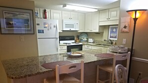 Una cocina con armarios blancos, microondas, estufa y pileta. Hay dos sillas y una mesa con un plato de comida.
