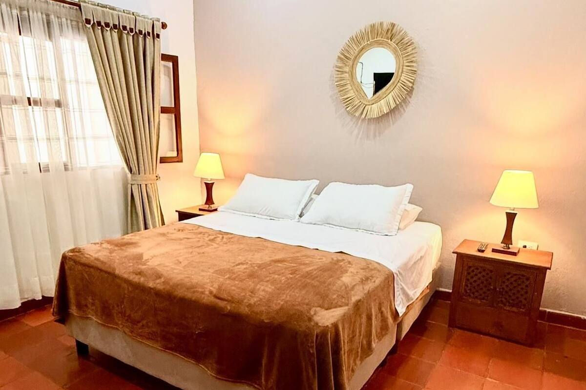 A hotel room with a bed, two bedside lamps, a mirror, and a window with curtains.