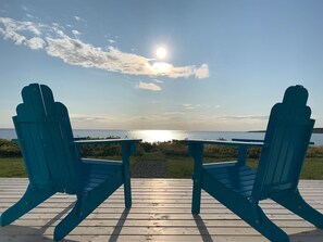 Two blue Adirondack chairs facing a wooden deck with a view of the ocean and a sunset. - Cabot`s Modern-Day Beach House (Chéticamp)