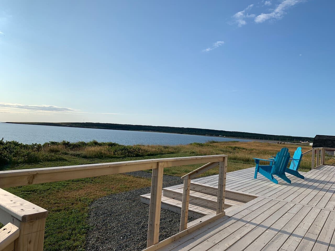 Eine Holzterrasse mit Geländer, zwei blaue Adirondack-Stühle und Blick auf ein Gewässer.