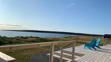 Eine Holzterrasse mit Geländer, zwei blaue Adirondack-Stühle und Blick auf ein Gewässer.