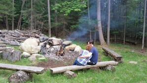 Un couple est assis près d’un feu de camp dans une zone boisée, entouré de bûches et de rochers.