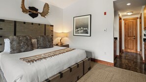 A bedroom with a large bed, a wooden headboard, a nightstand, a lamp, a framed picture, and a wooden door leading to a kitchen. - #1534 | Studio | Gondolier | 5Th floor | Mountain & Village Views (Stowe)