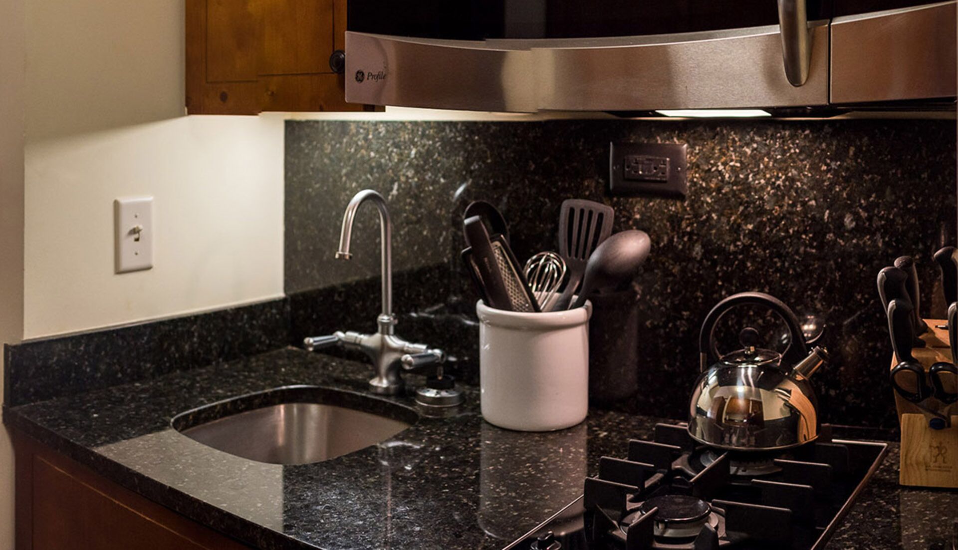 A kitchen with a stainless steel sink, a kettle, and a utensil holder.