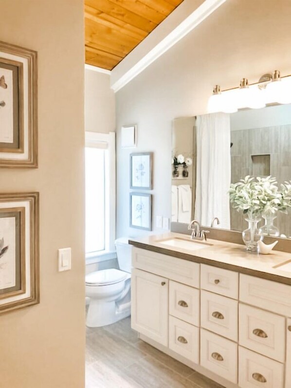A bathroom with a large mirror, a sink, and a toilet. There are framed pictures on the wall and a window with curtains.