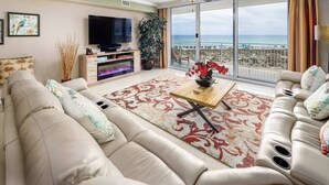 A living room with a sofa, a TV, a fireplace, a balcony with a view of the sea, and a ceiling fan.