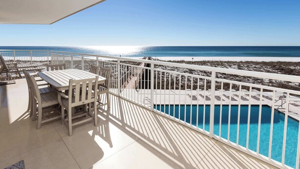 A balcony with a pool, white furniture, and a view of the beach and ocean.