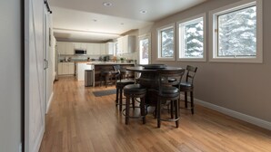 A modern kitchen with wooden flooring, a dining area with a round table and bar stools, and large windows with a view of snow-covered trees. - Beautiful Home in McCall w/ family & living room and large backyard w/ firepit (Mccall)