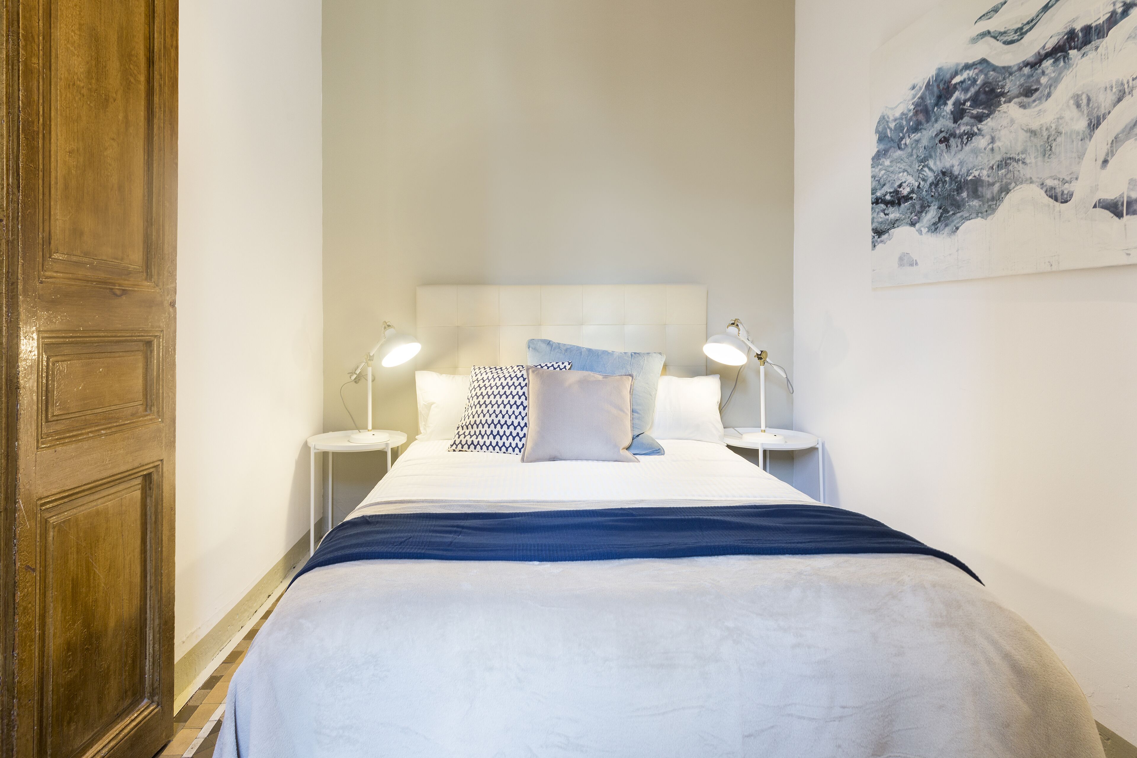 Een net opgemaakt bed met een wit hoofdbord, twee bedlampen en een blauw kussen.
