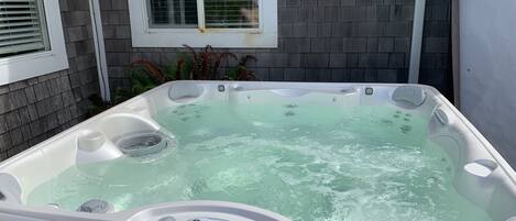 A hot tub with a control panel, set against a house exterior with a window and plants.