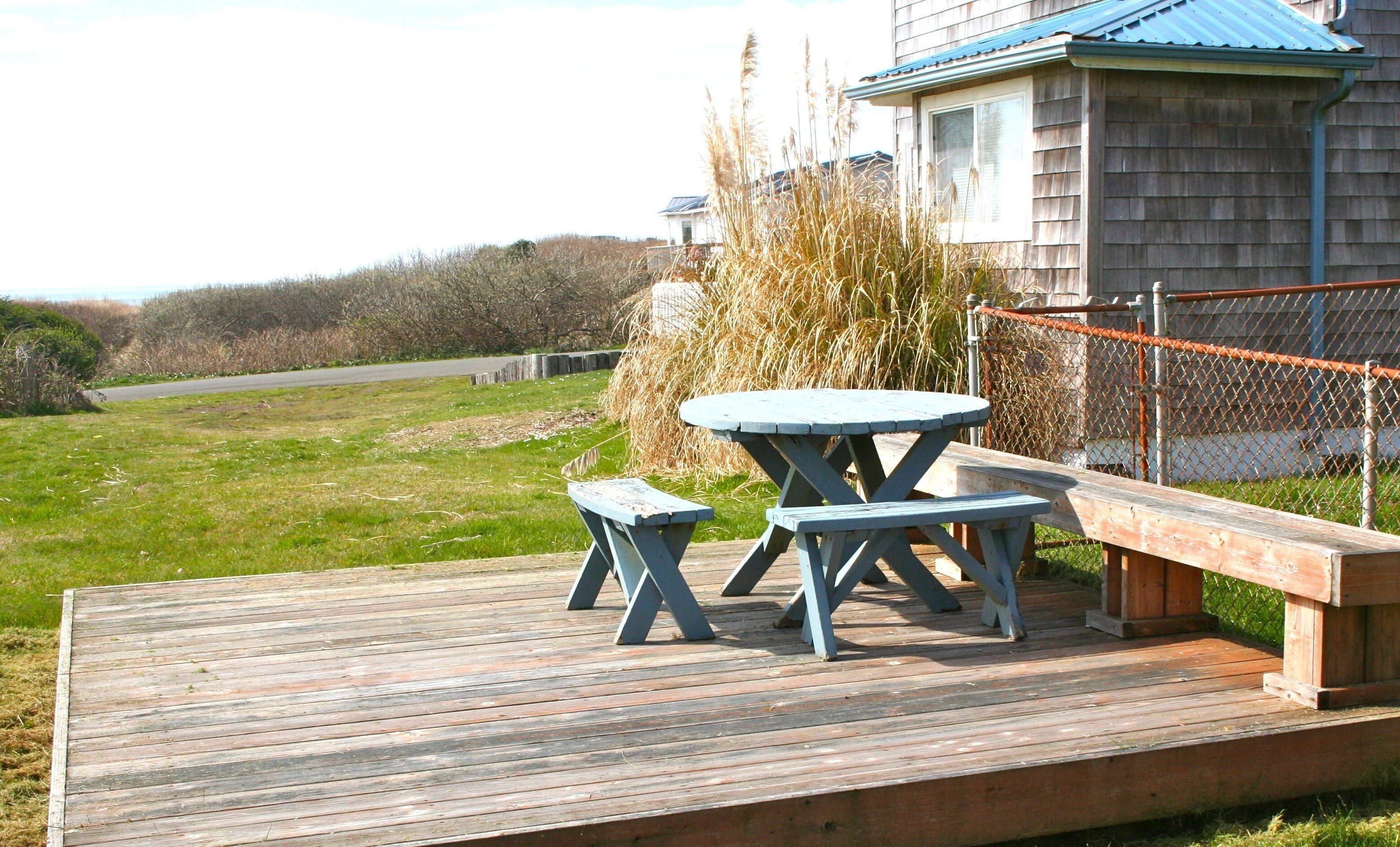 A wooden deck with a picnic table and benches, surrounded by a chain-link fence and a building with a blue roof.