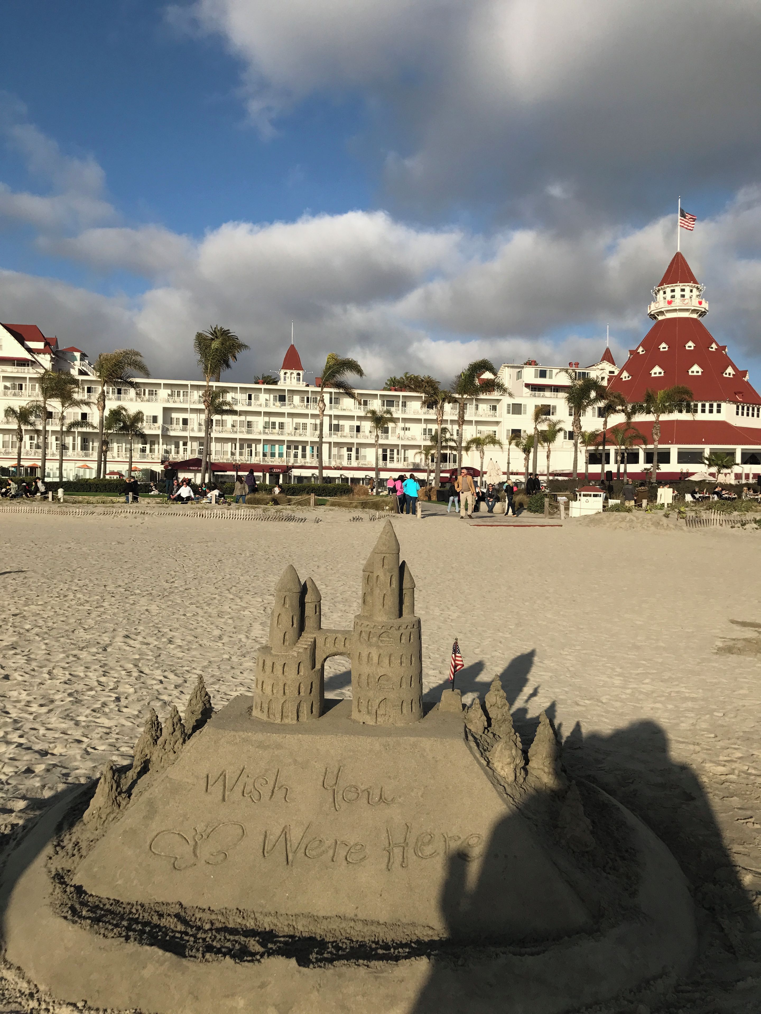 Una spiaggia con un castello di sabbia e un messaggio, un hotel con diversi piani e balconi, e una torre con il tetto rosso.