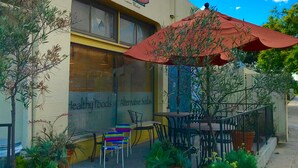 A storefront with a sign reading "TRAXS MARKETPLACE" and "Healthy Foods Alternative Sodas". There are outdoor tables and chairs, and potted plants around the entrance.